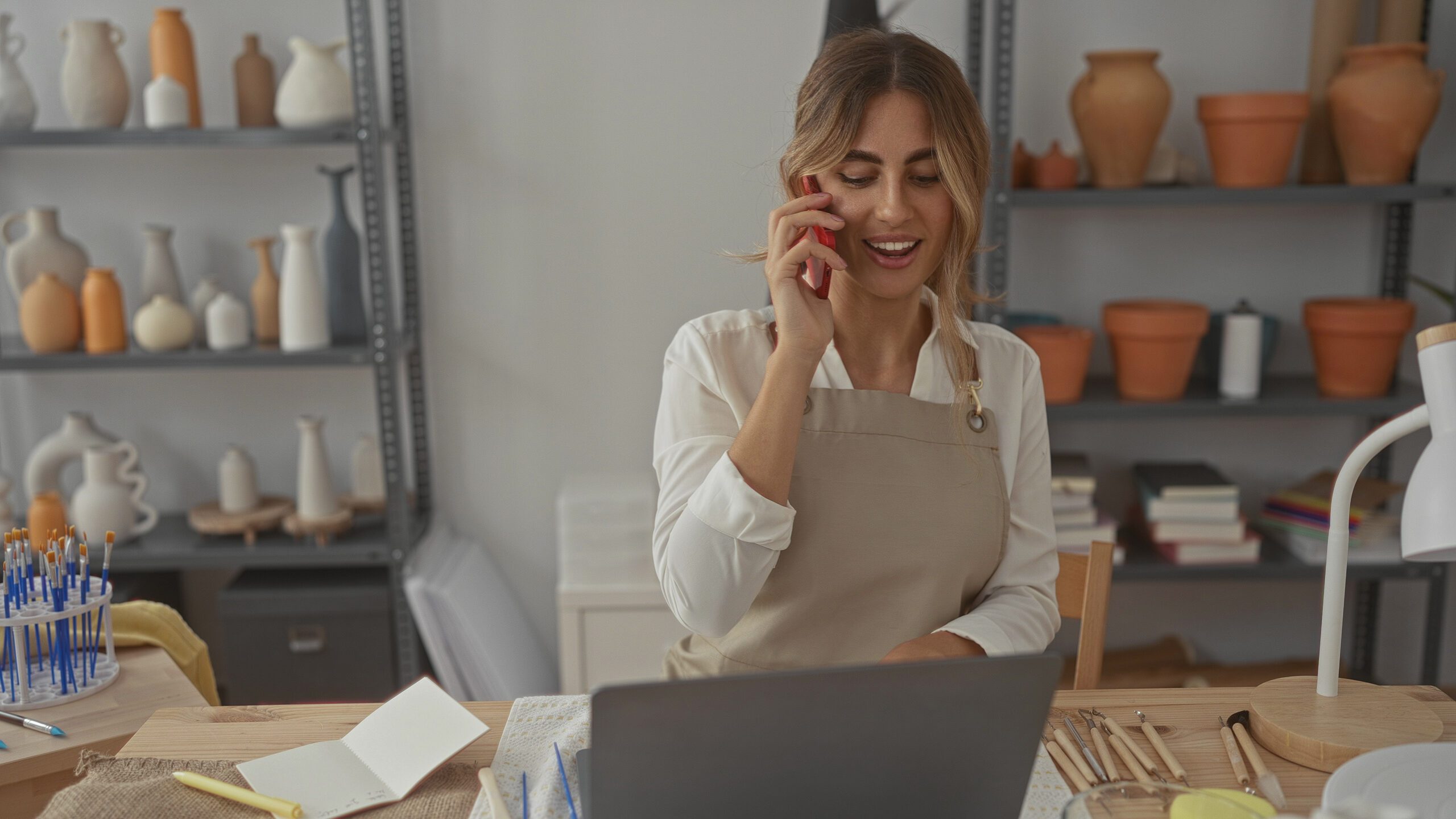 Young woman holding phone to ear in pottery studio while checking orders and reaching for clay tools; creativity focus serenity.