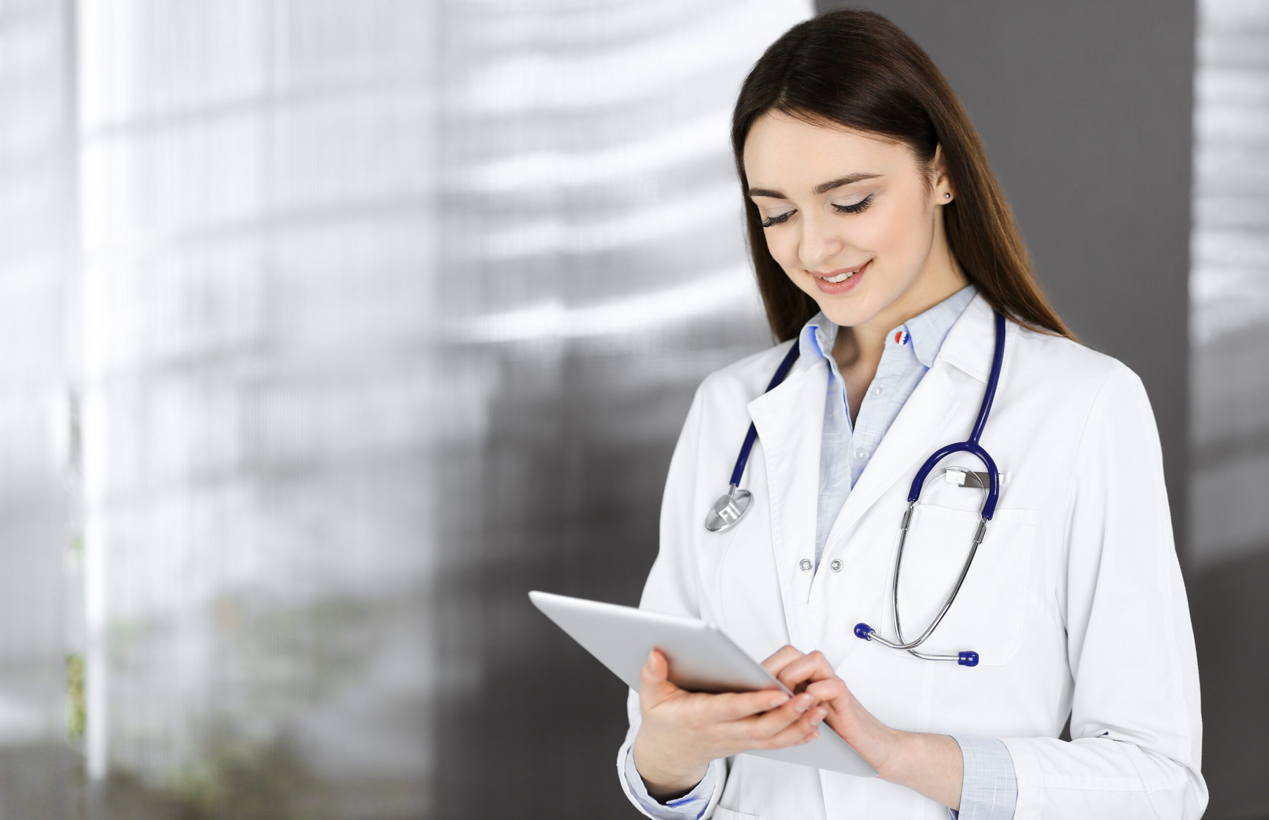 Cheerful intelligent woman-doctor is holding a tablet computer in her hands, while she is standing in a clinic. Portrait of a friendly physician woman at work. Perfect medical service in a hospital. Medicine concept