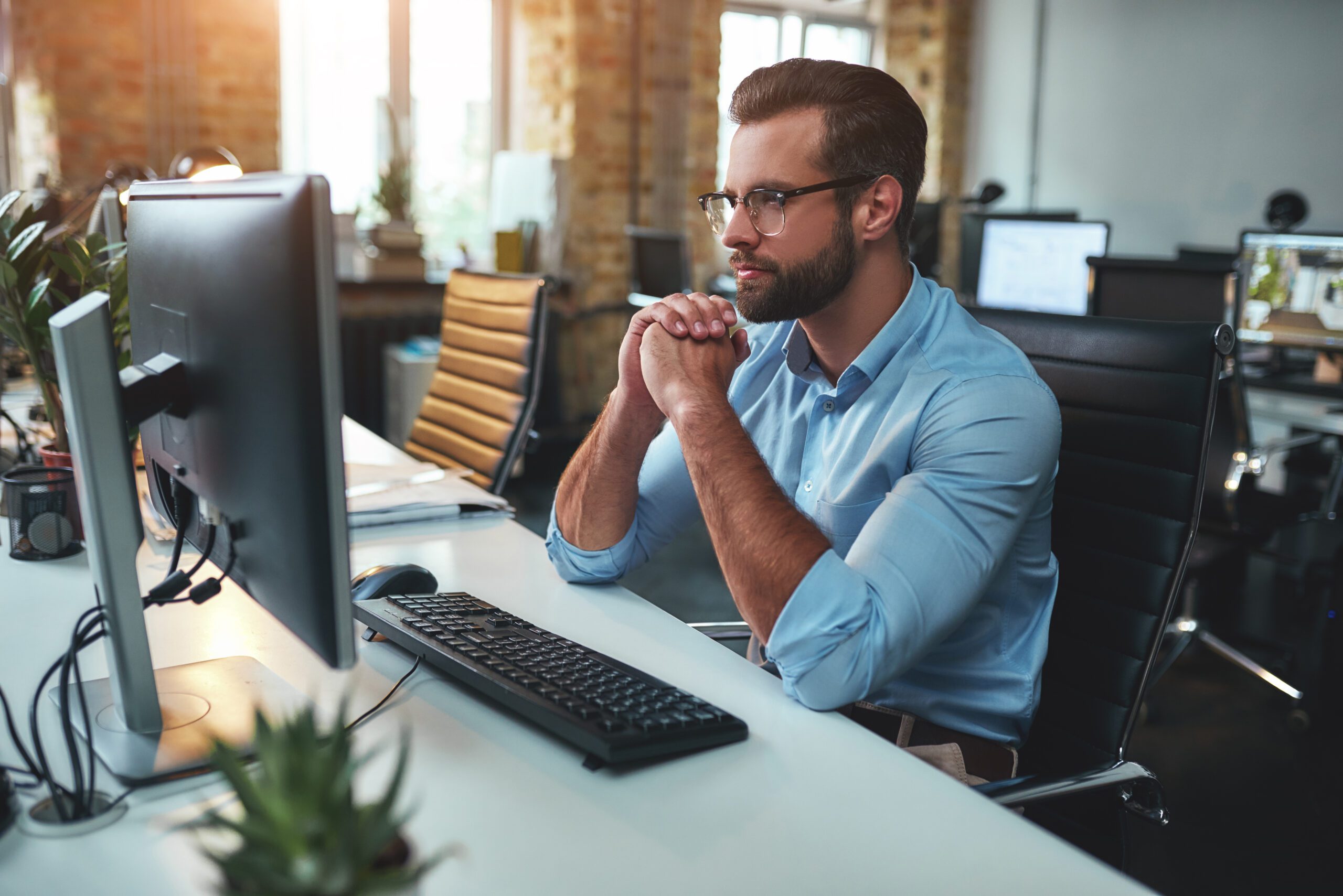 Thinking. Side view of young bearded man in eyeglasses and formal wear working on computer and keeping palms pressed together while sitting in the modern office