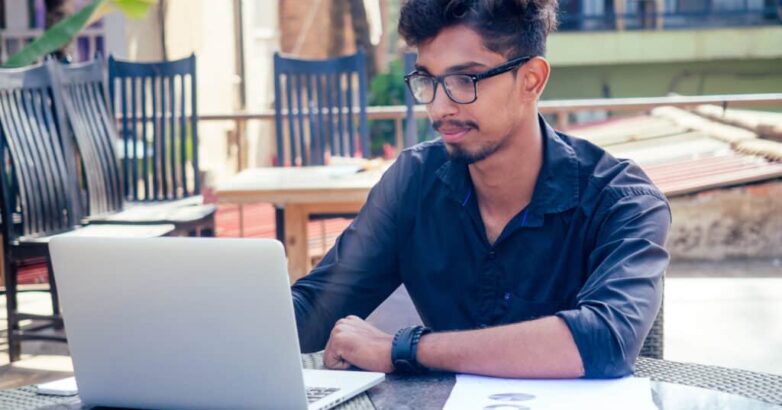 handsome and successful indian man in a stylish well-dressed freelancer working with a laptop on the beach.freelance and remote work.businessman student in a summer cafe on the shore of india ocean