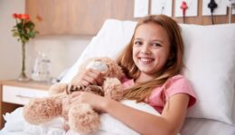 Portrait Of Girl Lying In Bed In Hospital Ward Hugging Teddy Bear