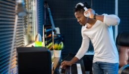 Young serious casual mixed-race man with headphones standing by computer screen