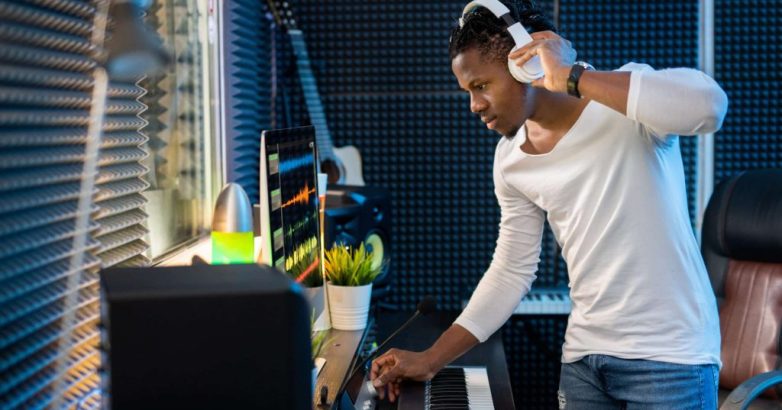 Young serious casual mixed-race man with headphones standing by computer screen