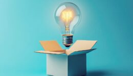 Glowing lightbulb emerging from open white cardboard box against turquoise backdrop, symbolizing breakthrough creativity
