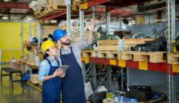 Portrait of two modern factory workers, man and woman, wearing hardhats doing inventory standing by tall shelves in warehouse pointing up, copy space