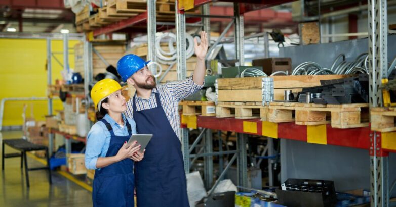 Portrait of two modern factory workers, man and woman, wearing hardhats doing inventory standing by tall shelves in warehouse pointing up, copy space