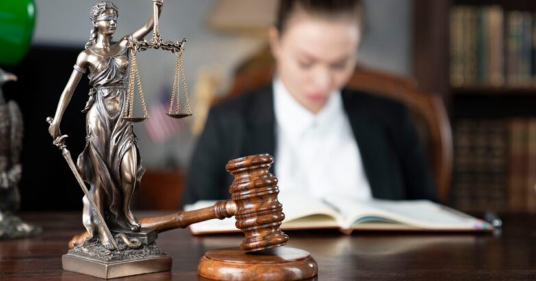 Young female lawyer during work in chamber. Gavel and Themis statue on the brown shining desk.