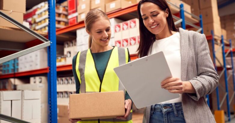 Female Team Leader With Clipboard In Warehouse Training Intern Standing By Shelves Female Team Leader With Clipboard In Warehouse Training Intern Standing By Shelves