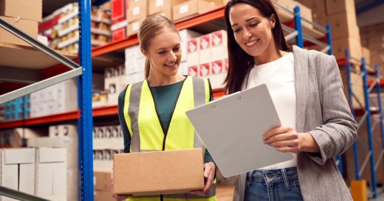 Female Team Leader With Clipboard In Warehouse Training Intern Standing By Shelves