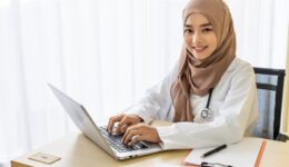 Confident Muslim female woman medical doctor sitting at desk, working typing with laptop computer in clinic hospital office; looking at camera