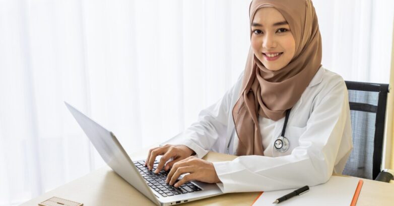 Confident Muslim female woman medical doctor sitting at desk, working typing with laptop computer in clinic hospital office; looking at camera Confident Muslim female woman medical doctor sitting at desk, working typing with laptop computer in clinic hospital office; looking at camera