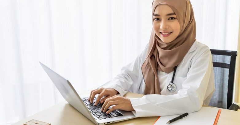 Confident Muslim female woman medical doctor sitting at desk, working typing with laptop computer in clinic hospital office; looking at camera