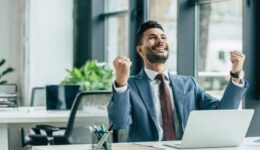 happy businessman looking up and showing winner gesture while sitting at workplace