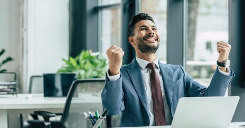 happy businessman looking up and showing winner gesture while sitting at workplace