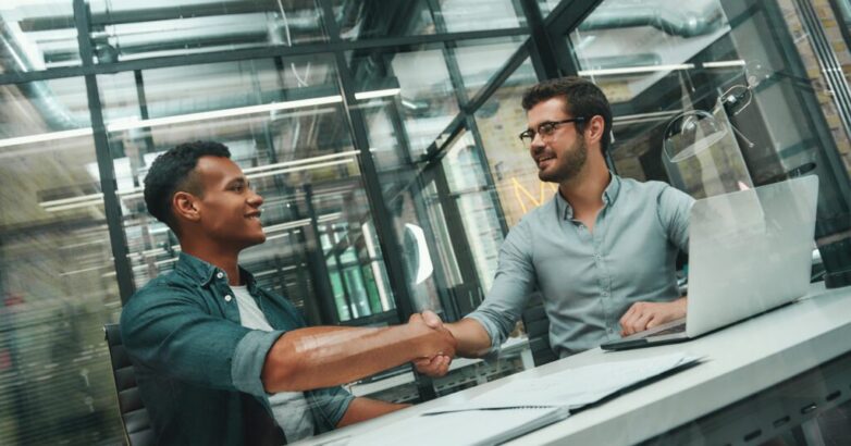 Welcome on board! Two young and handsome men shaking hands and smiling while working in modern office