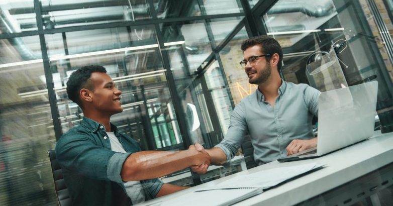 Welcome on board! Two young and handsome men shaking hands and smiling while working in modern office